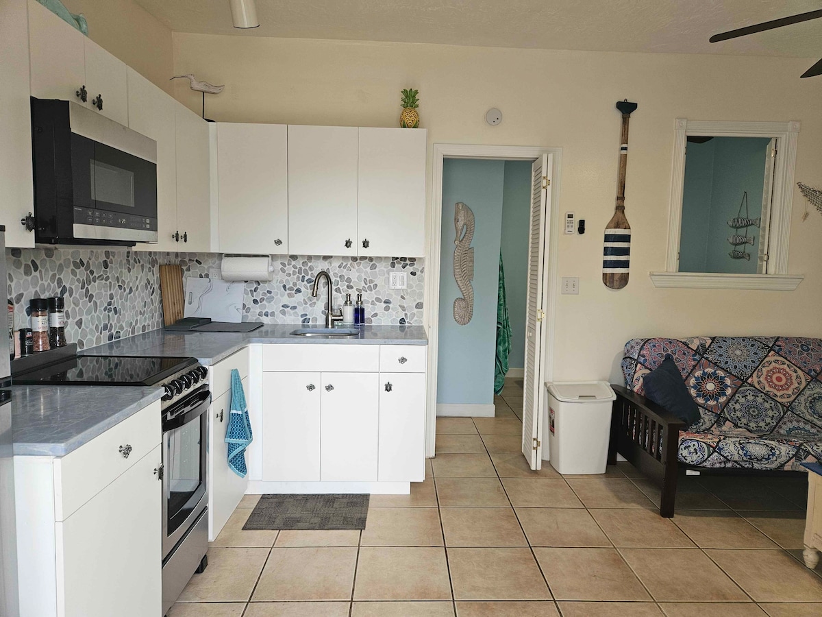 The kitchen area features modern appliances, including a microwave and stove, set against a decorative backsplash. White cabinetry offers ample storage, and a small multi-colored futon couch is positioned nearby. Tiled flooring enhances the open feel of this functional space.