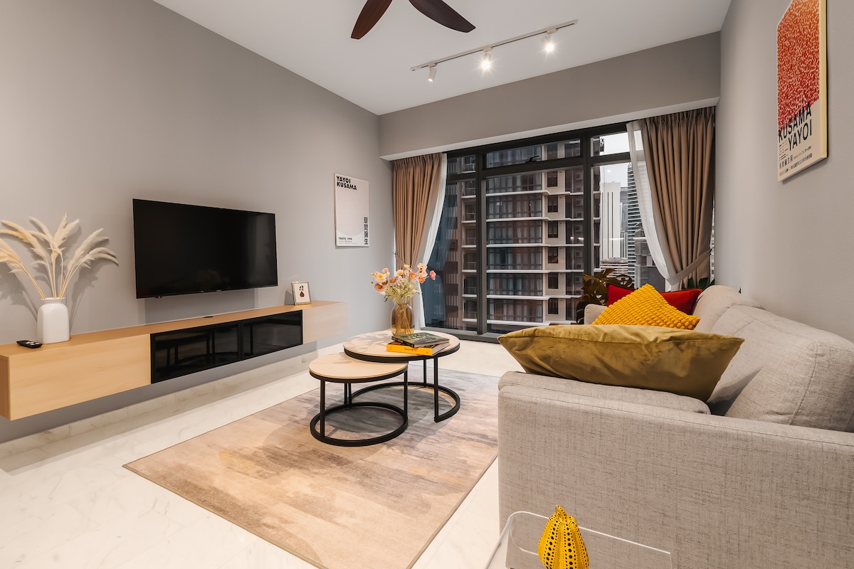 The living area features a light-colored sofa with throw pillows, complemented by a sleek coffee table. A flat-screen TV is mounted on the wall, and natural light filters through large windows, covered with sheer curtains. Decorative plants add a touch of warmth to the space.