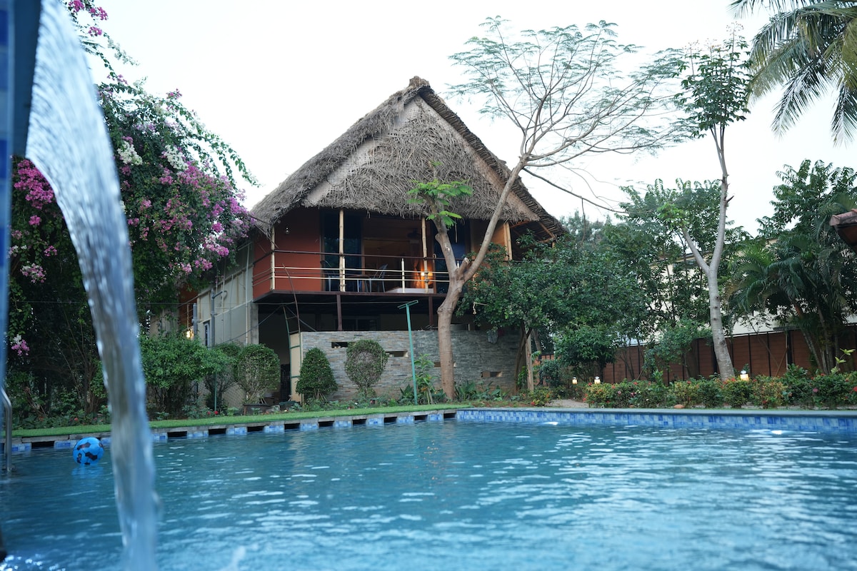 A thatch-roof house is visible in the background, surrounded by lush greenery and flowering plants. In the foreground, a pool reflects the sky, while a gentle waterfall feature adds a calming sound. The overall setting conveys a tranquil and natural environment.