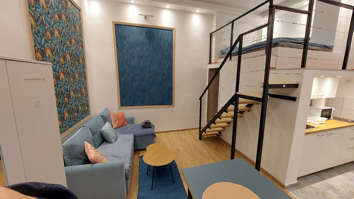A modern living area features a light-blue couch and two coffee tables, providing a comfortable seating arrangement. A decorated wall art piece adds a splash of color, while a staircase leads to a mezzanine with a spacious bed. Warm lighting highlights the wooden flooring.