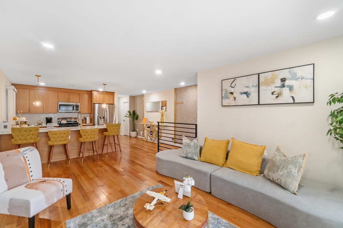 An open-concept living space is highlighted by warm wood flooring and modern furnishings. A cozy sofa adorned with yellow cushions faces a round coffee table. The kitchen features a breakfast bar with four stools, and artwork decorates the wall to add a contemporary touch.