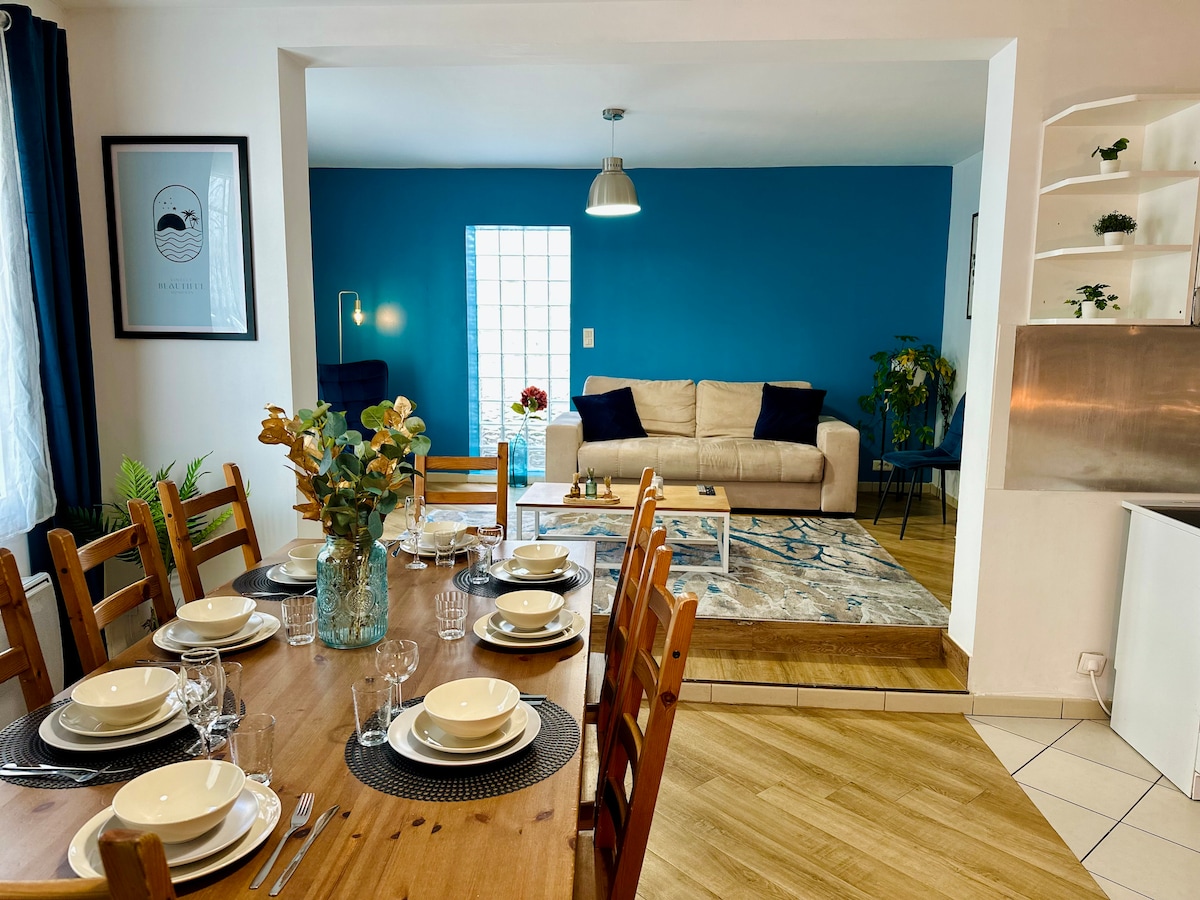 A spacious living and dining area is visible, featuring a wooden dining table set for eight. The modern sofa is placed against a vibrant blue wall, complemented by a rug and decorative plants. Natural light enters through a window, enhancing the inviting ambiance.