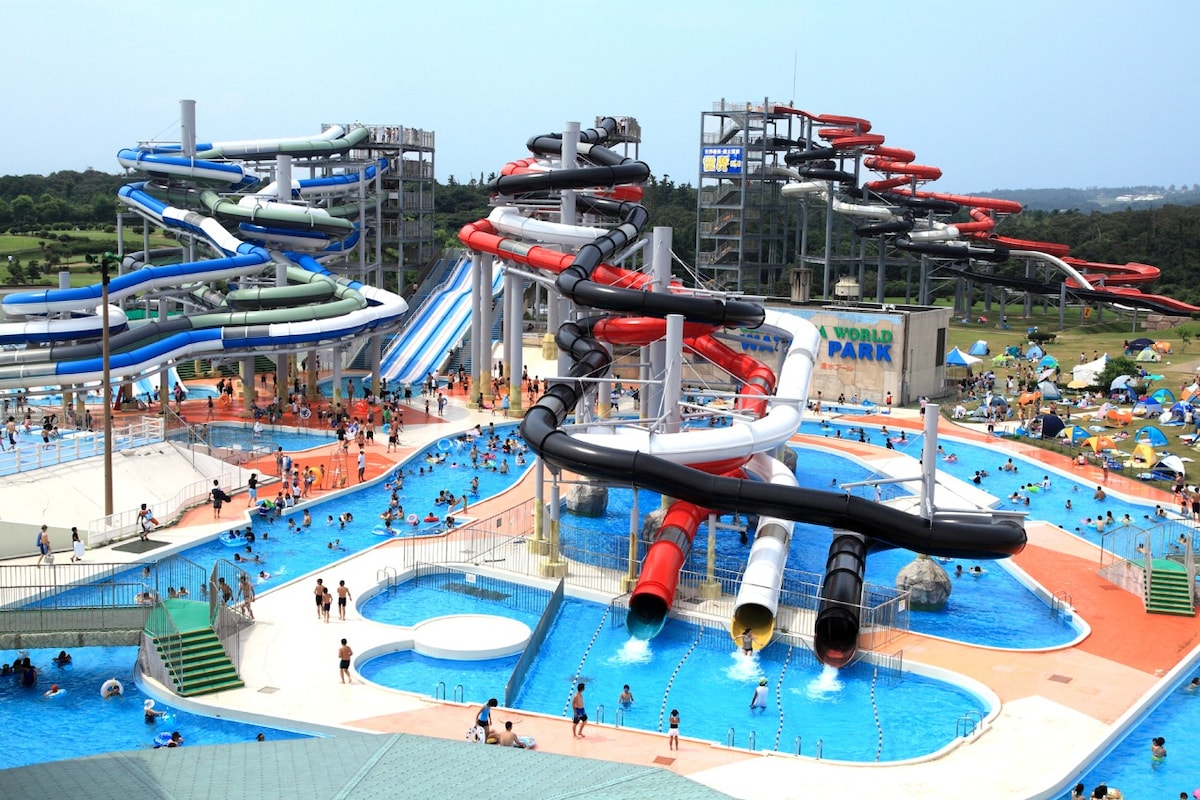 A large water park features a variety of colorful water slides twisting down into a swimming pool area. Families and individuals are enjoying the water, while grassy areas with tents for relaxation are visible in the background. Sunlight reflects off the water, creating a lively atmosphere.