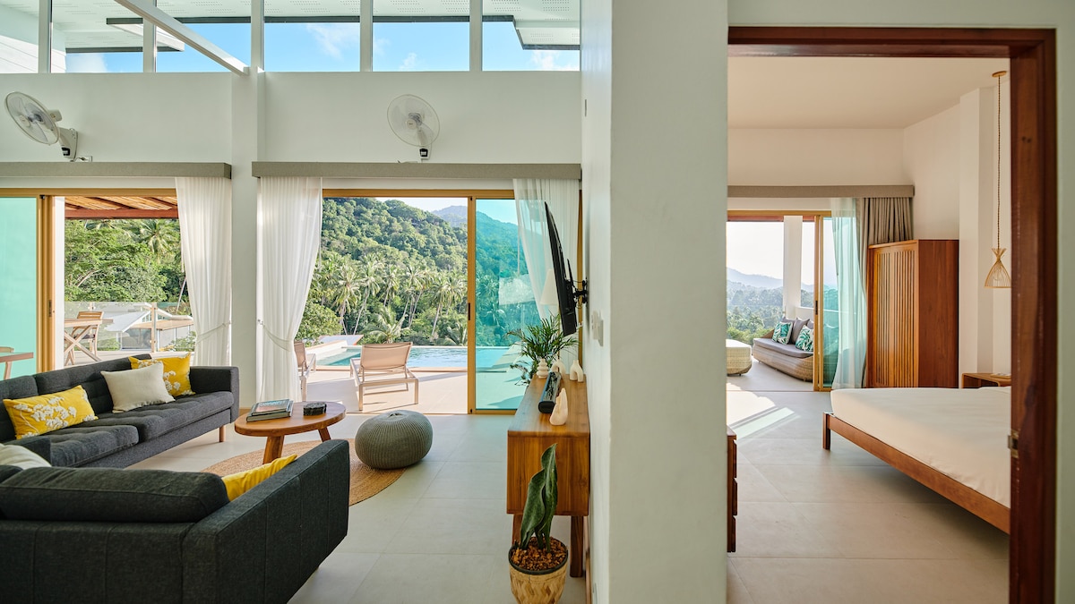 An open-plan living area connects to the outdoor terrace, featuring a comfortable gray sofa, a wooden television unit, and large sliding glass doors that reveal sea views. Nearby, a bright bedroom is adorned with natural light, showcasing a spacious bed and a cozy seating area.