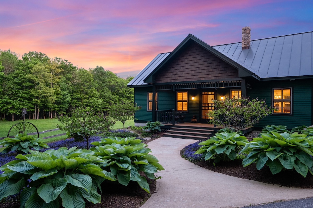 The exterior of a custom-built home is showcased against a vibrant sunset. Lush green foliage and blooming hostas frame a winding path leading to wooden doors. The home features a dark exterior with large windows, allowing natural light to illuminate the interior.