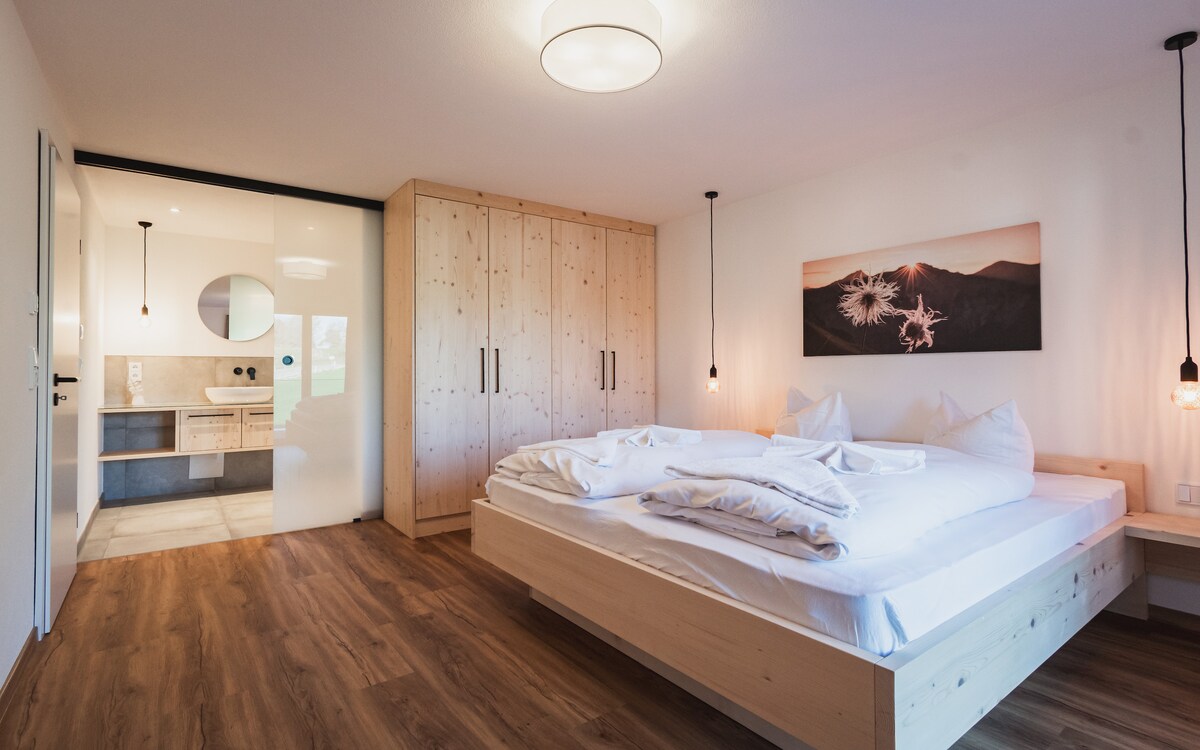 A modern bedroom features a spacious bed with crisp, white bedding. Light wooden elements create a natural look. Two pendant lights hang over the bed, providing soft illumination. A stylish photograph adorns the wall. A glass door leads to a well-appointed bathroom.