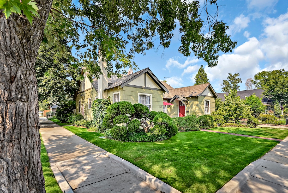 The exterior of a vintage home features charming green siding, surrounded by manicured lawns and well-kept shrubs. A tree stands prominently in the foreground, framing the property, while a path leads invitingly towards the entrance.