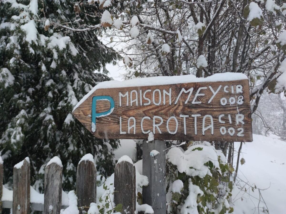 A wooden directional sign covered in snow points towards "MAISON MEY" and "LA CROTTA," with the letters painted in light blue against the weathered wood. Surrounding greenery is dusted with a light layer of snow, creating a serene winter scene.
