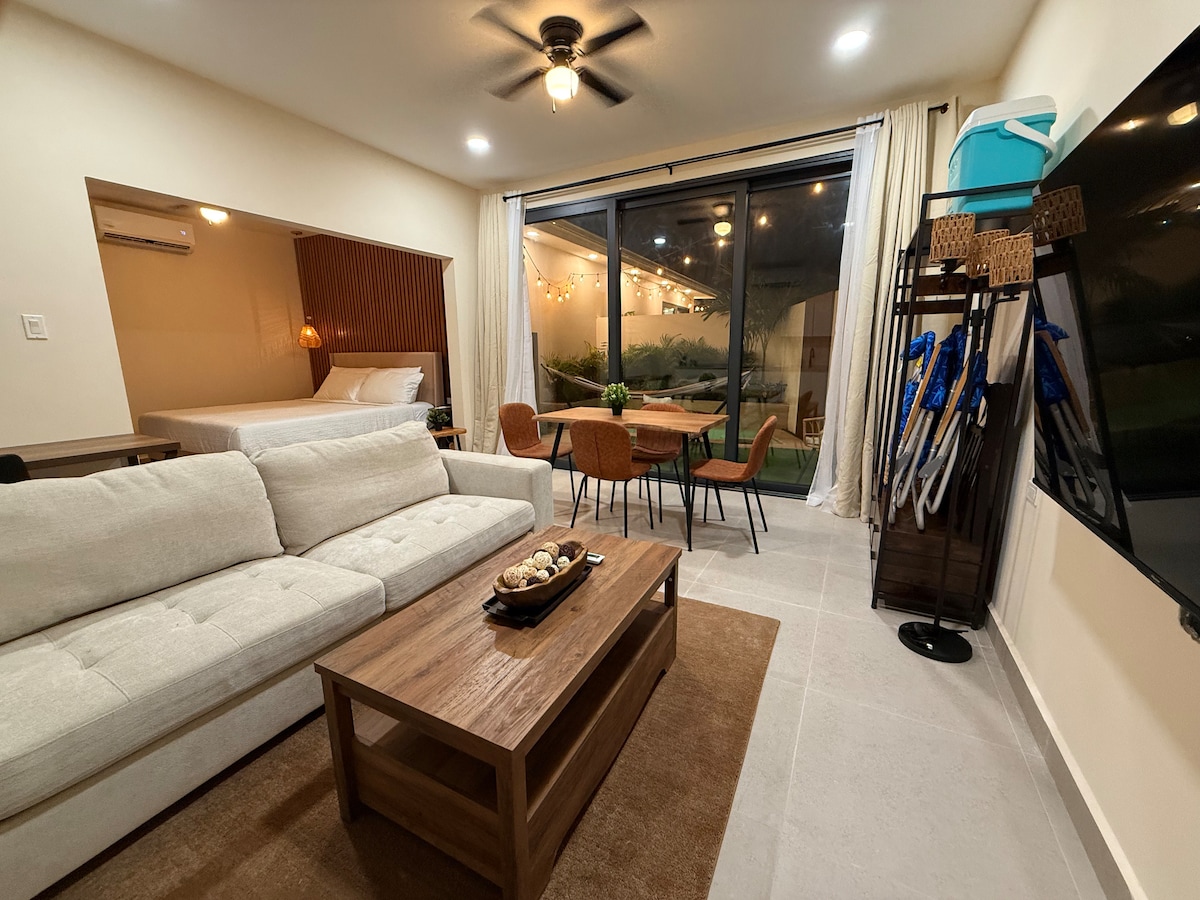 A modern studio living area features a light-colored sofa and a wooden coffee table placed on a textured rug. A dining set with four chairs is positioned beside large glass doors, revealing a view of the private plunge pool and outdoor greenery.