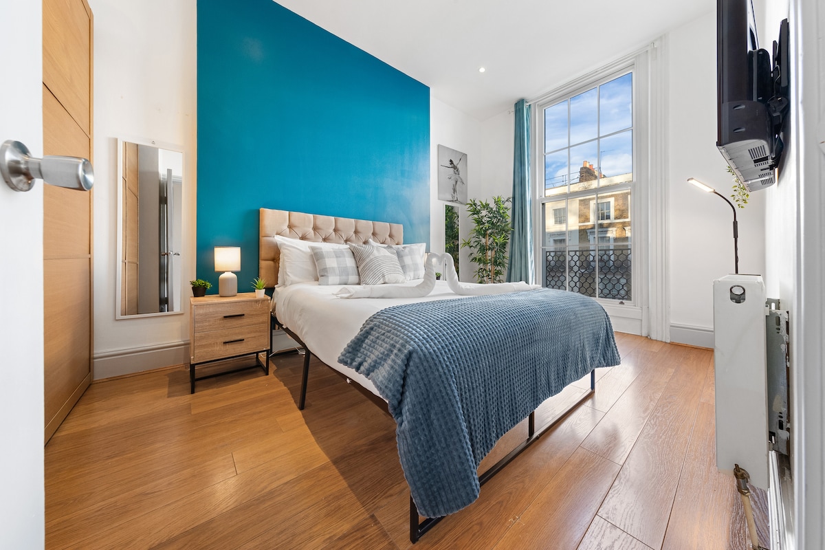 A bright bedroom features a spacious bed with a light grey tufted headboard and crisp linens. A large window provides abundant natural light, complemented by light curtains. A stylish tall mirror stands beside a wooden nightstand with a small plant. A blue accent wall adds a modern touch.
