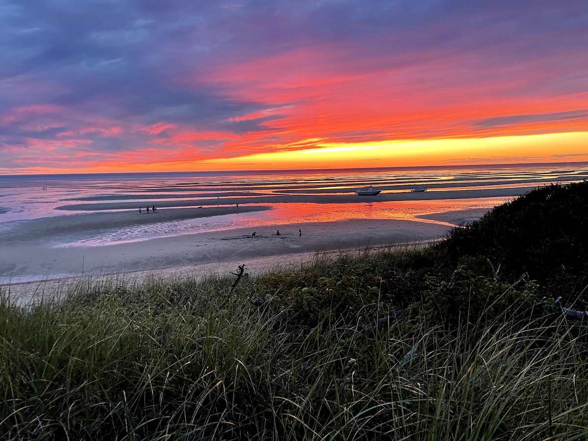 A vibrant sunset paints the sky in deep oranges and purples above a tranquil beach scene. Silhouetted figures can be seen walking along the wet sand, while distant boats rest on the shallow waters. Soft grass borders the foreground, enhancing the natural landscape.
