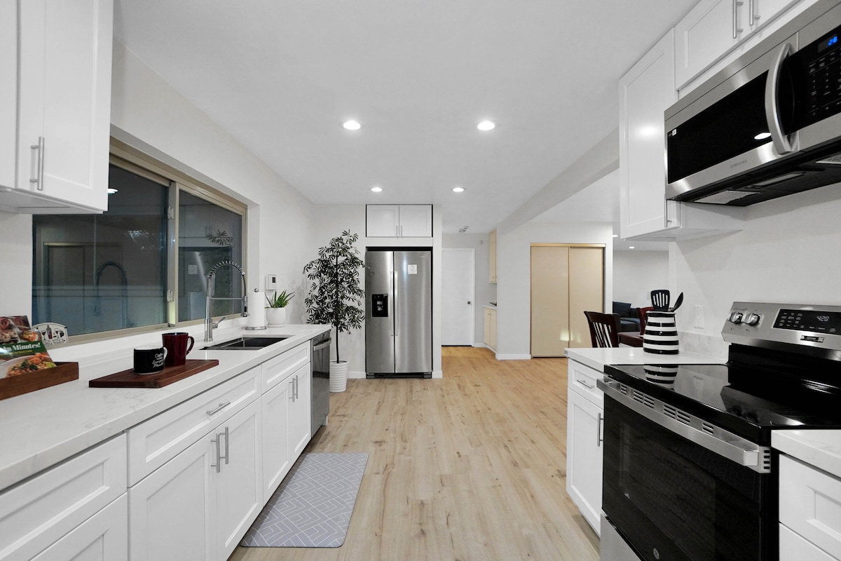A fully equipped kitchen features white cabinetry and modern stainless steel appliances. A large window offers natural light, while light-colored flooring adds to the bright atmosphere. Counter space is available for meal preparation, complemented by a rug and small decorative items.