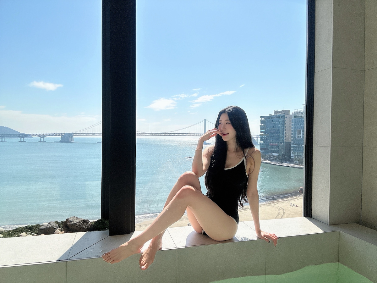 A figure is seated by a large window overlooking a panoramic ocean view of Gwangandaegyo Bridge and the shoreline. Natural light fills the space, enhancing the serene coastal atmosphere. The interior features a luxurious jacuzzi, creating a peaceful spot for relaxation.