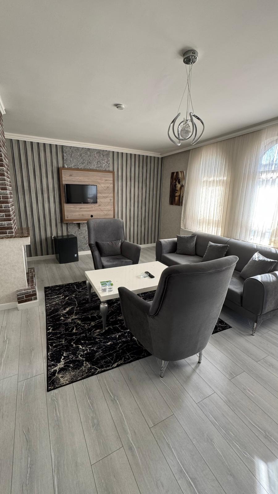 A modern living area features two comfortable armchairs and a cozy sofa surrounding a light coffee table. A flat-screen TV is mounted above a decorative accent wall. Natural light filters through sheer curtains, illuminating the neutral color palette and patterned rug.