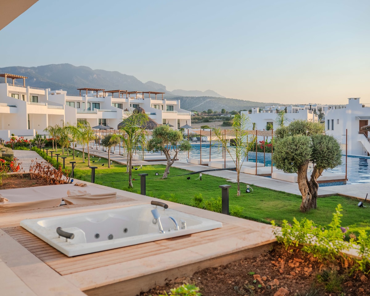 A spacious terrace is shown featuring a white jacuzzi, surrounded by a deck and lush greenery. In the background, a well-kept garden and multiple residential buildings are visible, along with a large pool area and distant mountains under a clear sky.