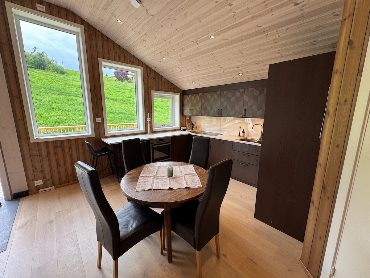 A modern kitchen area is depicted with wood paneling throughout. A round table surrounded by four dark leather chairs sits at the center. The kitchen features sleek cabinetry, and sunlight streams in from three large windows, creating a bright and inviting workspace.