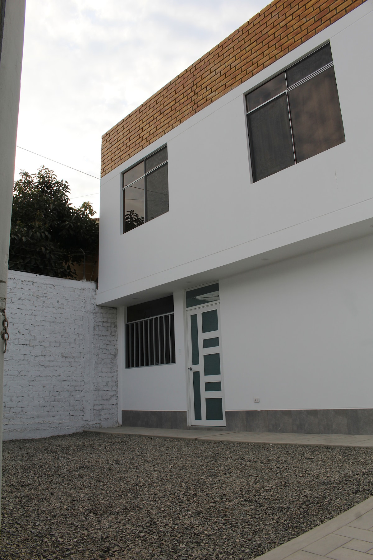 The exterior view showcases a two-story building with a clean white facade and large windows allowing natural light. The entrance features a modern door with multiple panels. A gravel area is visible in the foreground, surrounded by a plain wall and greenery in the background.