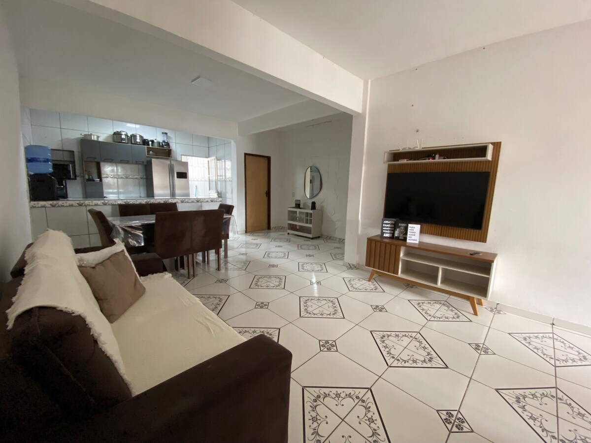 An open living area features a comfortable brown sofa with accent pillows, positioned next to a stylish dining set. The tiled floor adds a modern touch, while a large flat-screen TV is mounted on the wall opposite. A well-equipped kitchen space is visible in the background.