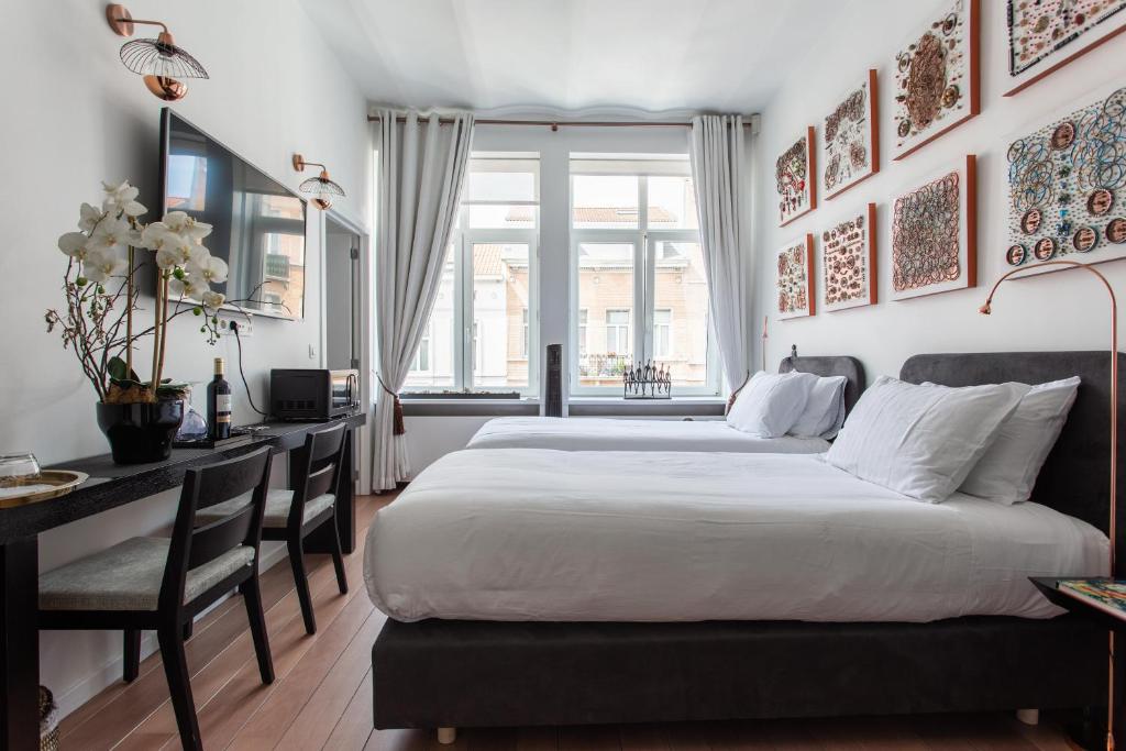 A well-lit room features two comfortable beds with white bedding. Artwork adorns the walls, and a flat-screen TV is mounted above a compact desk. Large windows allow natural light to fill the space, while light curtains frame the view of the outside.