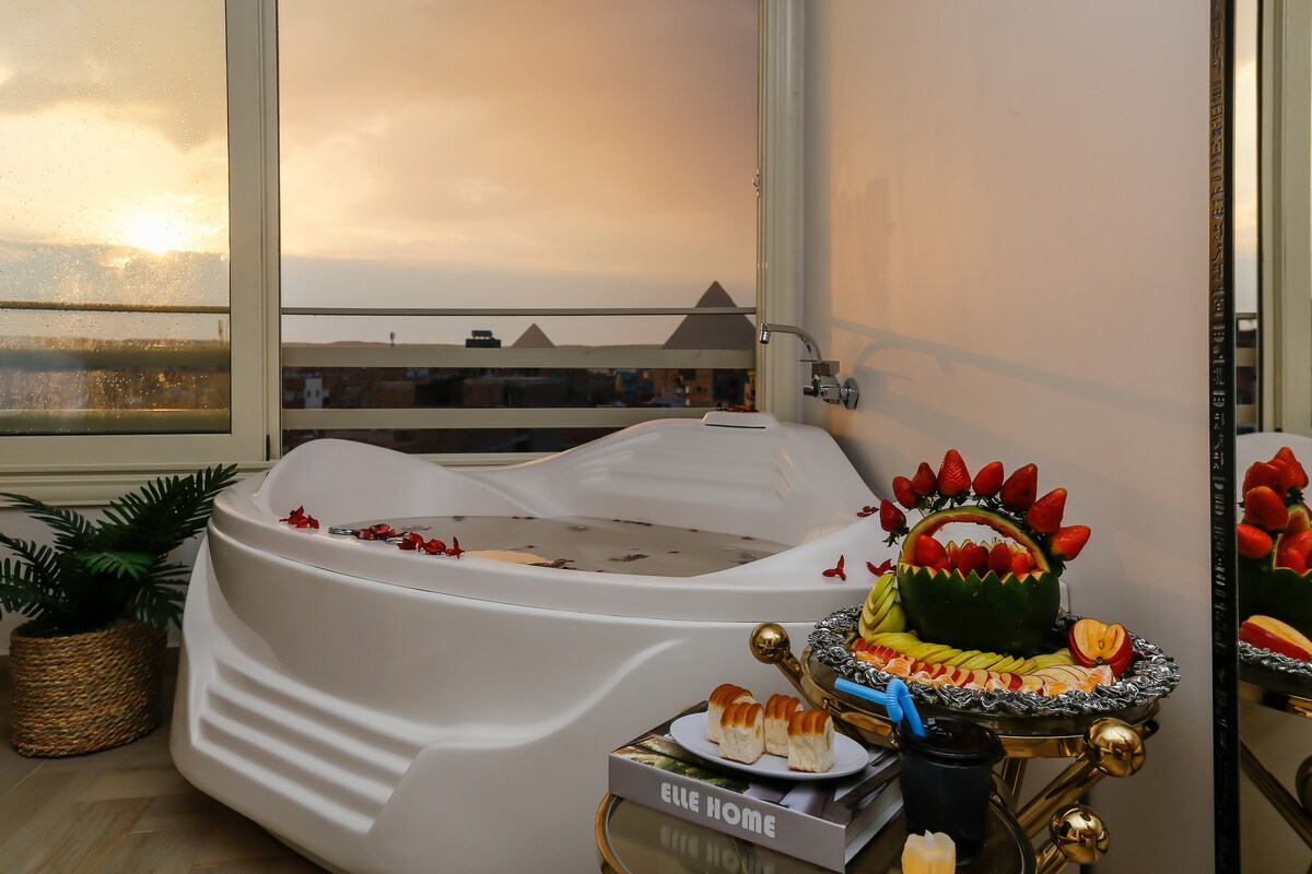 A luxurious Jacuzzi is positioned near a window, offering a scenic view. The tub features petals floating on the water's surface. A decorative tray holds a variety of fruits and pastries, enhancing the inviting appeal. Soft lighting creates a serene ambiance.