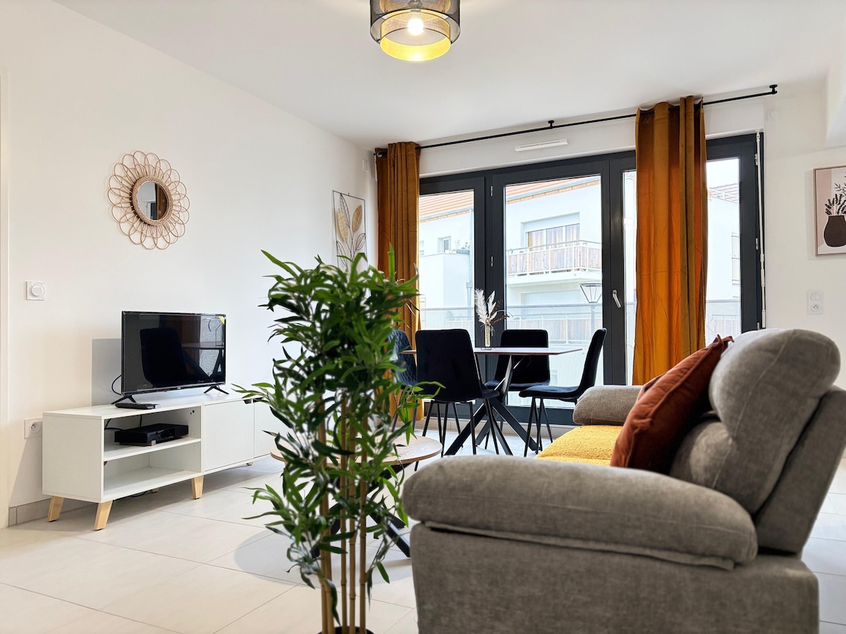 A modern living space is showcased with a comfortable gray sofa positioned near a small round table for four. Large windows allow ample natural light, with sheer curtains enhancing the brightness. A flat-screen TV and decorative accents add to the inviting atmosphere.