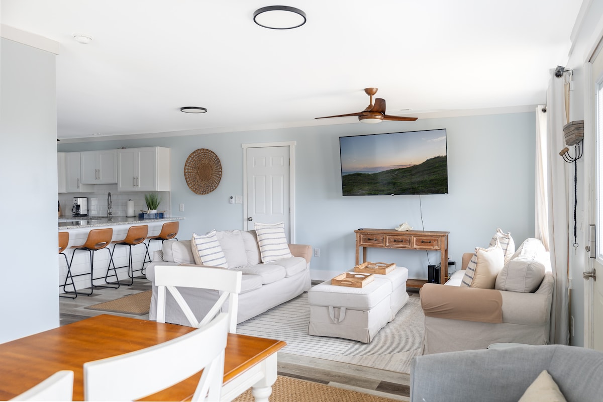 A modern living space features light-colored furnishings, including a sectional sofa and accent chairs. A large flat-screen TV is mounted on the wall, while a wooden console table adds functionality. The open layout connects to a well-equipped kitchen with bar seating.