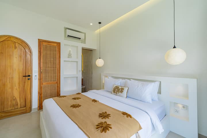 غرفة نوم مشرقة ومتجددة الهواء تتميز بنافذة كبيرة مقوسة مع إطلالات خصبة، وسرير أبيض مريح مع رميات زخرفية، ومرآة كاملة الطول، ومكتب خشبي بسيط في الزاوية. يضيف الضوء المعلق الحديث لمسة دافئة.