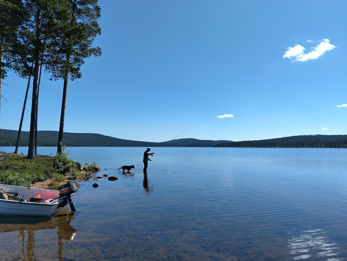A serene lakeside scene is depicted, featuring a person fishing along the shore. Calm waters reflect the clear blue sky, while distant mountains provide a scenic backdrop. A small boat is moored nearby, contributing to the tranquil atmosphere of the natural surroundings.