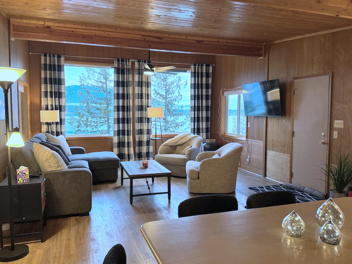 A cozy living area features comfortable seating with a mix of gray sofas and accent chairs. Large windows frame scenic lake views, complemented by plaid curtains. A coffee table is positioned centrally, while a flat-screen TV is mounted on the wall, enhancing the inviting space.