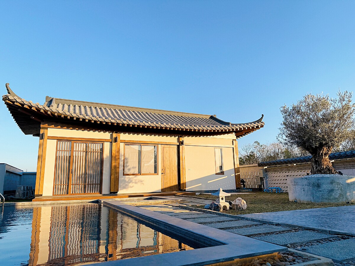A traditionally designed building features a curved roof and wooden accents, reflecting a unique architectural style. The structure is complemented by a tranquil pool, with clear water mirroring the bright blue sky and neatly arranged stone pathways leading to the entrance.