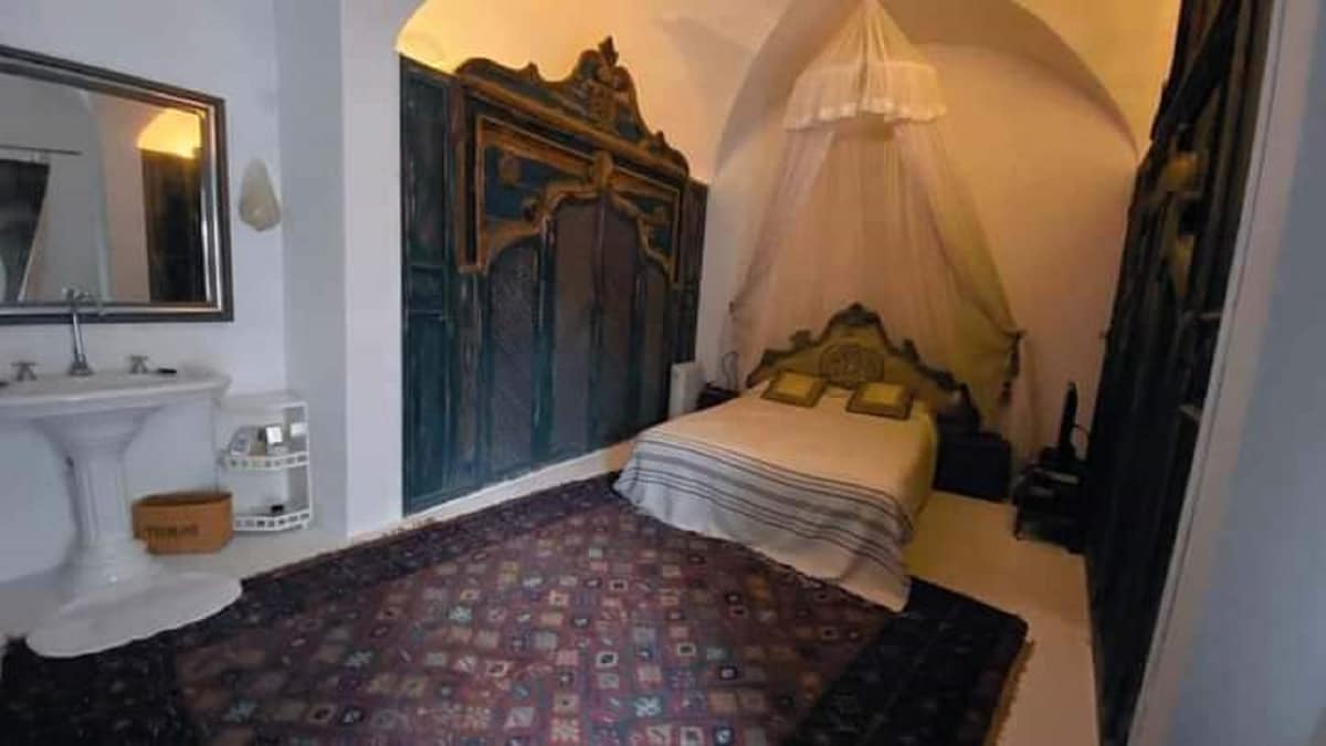 A spacious bedroom features a large bed adorned with patterned linens. Decorative wooden cabinetry frames the space, while a traditional area rug adds warmth. A simple washstand is positioned nearby, and soft lighting creates an intimate setting.