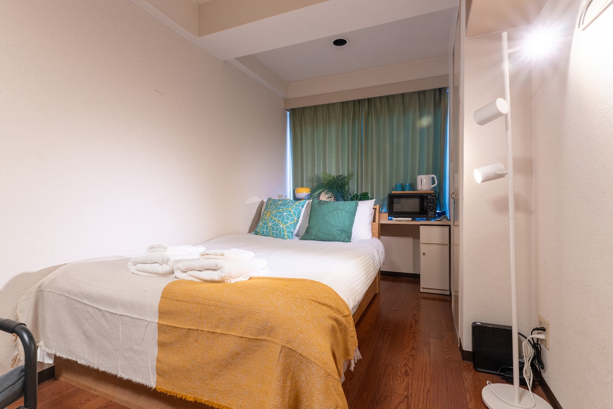 A comfortable bedroom features a spacious bed adorned with decorative pillows and a soft throw blanket. Natural light filters through the green curtains, illuminating a small table and a microwave at the corner. Fresh towels are arranged neatly on the bed.