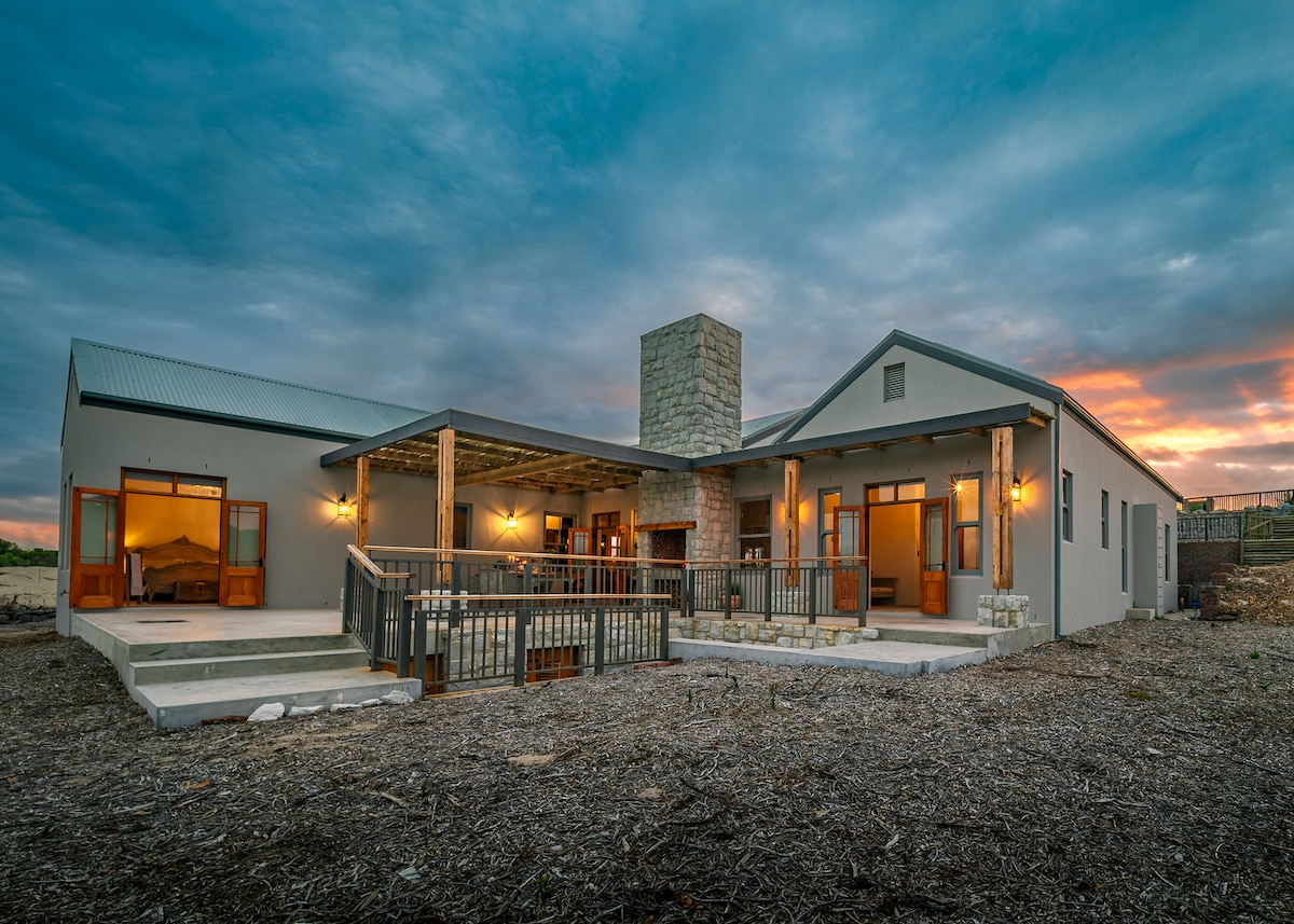 A spacious home exterior is highlighted at dusk, featuring a stone chimney and wooden beams on the porch. Large windows allow for natural light, while the surrounding landscape is marked by gravel and natural elements, blending the structure harmoniously with its environment.