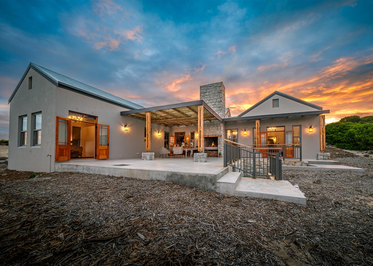 A modern building with a stone fireplace is highlighted against a dramatic sunset sky. The exterior features large glass doors that open to a spacious patio, complemented by rustic wooden beams and thoughtfully placed outdoor seating.