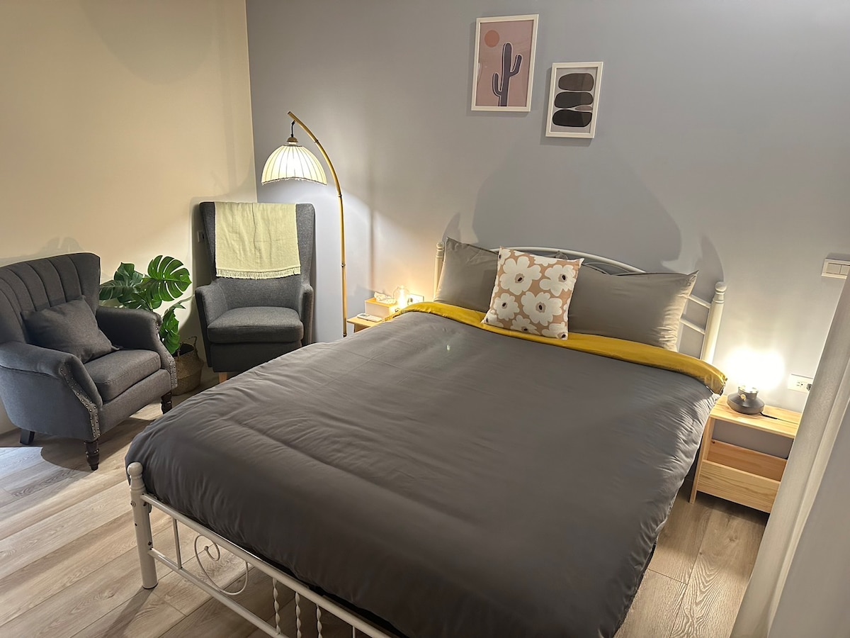 A cozy sleeping area features a spacious bed with a dark gray comforter and a yellow accent blanket. Two armchairs are positioned nearby, one adorned with a light green throw. Soft lighting from a lamp creates a calm ambiance, complemented by minimal wall art.