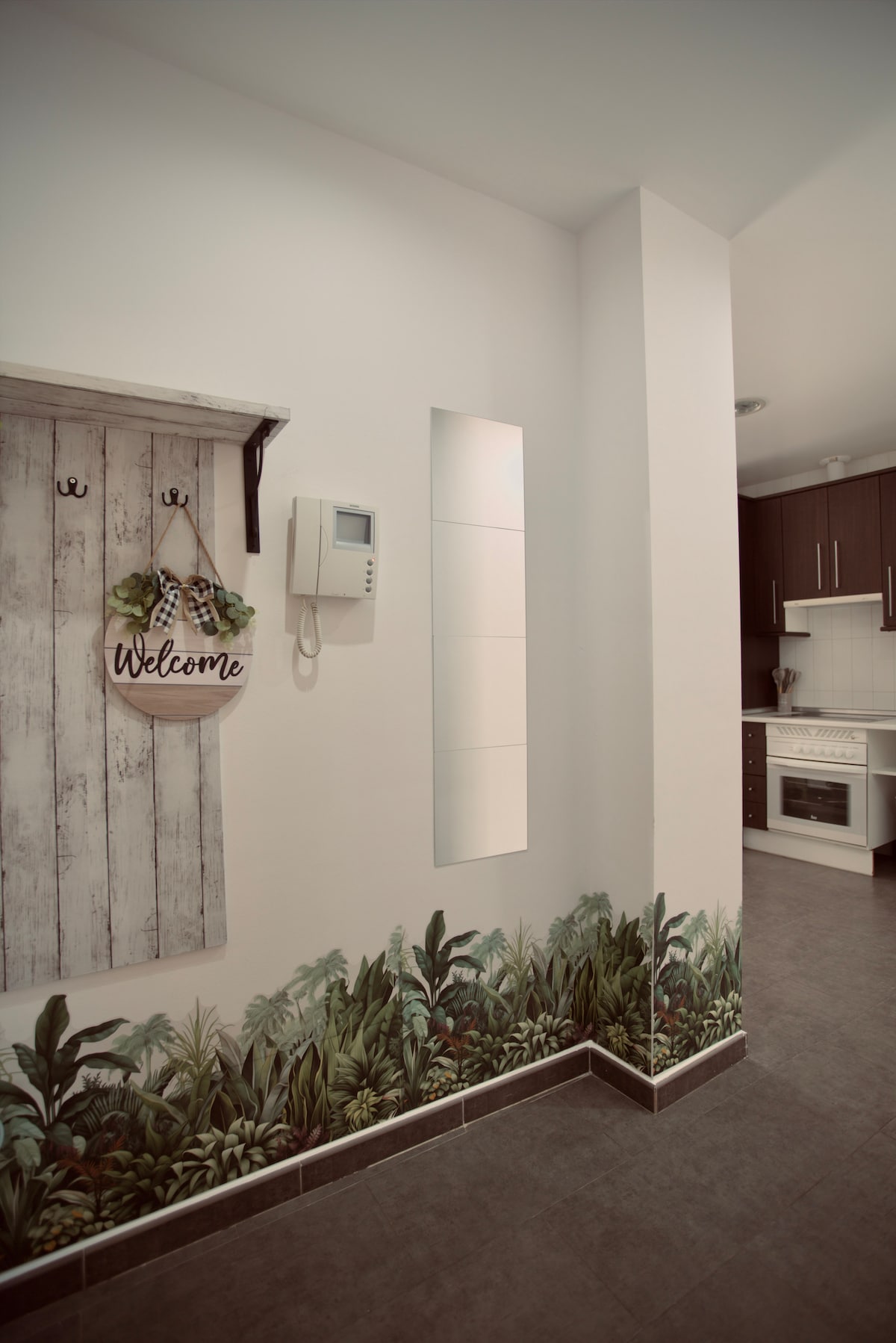 A modern entryway is depicted, featuring a wall-mounted welcome sign among decorative hooks. A soothing mural of greenery lines the base of the wall, contrasting with the sleek tile flooring. A doorway leads to a stylish kitchen area in the background.