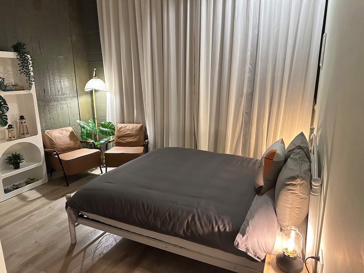 A cozy bedroom is highlighted by a spacious bed with soft bedding. Two comfortable armchairs are positioned beside a lamp, creating an inviting reading nook. Light curtain panels soften the atmosphere, and decorative plants add a touch of greenery to the space.