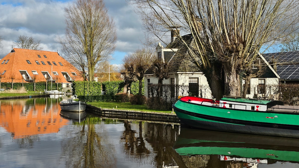 A serene view of a marina showcases moored boats beside a tranquil canal. Reflections of nearby trees and structures are visible on the water's surface, creating a harmonious atmosphere. A nearby building features a distinctive orange roof and solar panels, contributing to the natural setting.