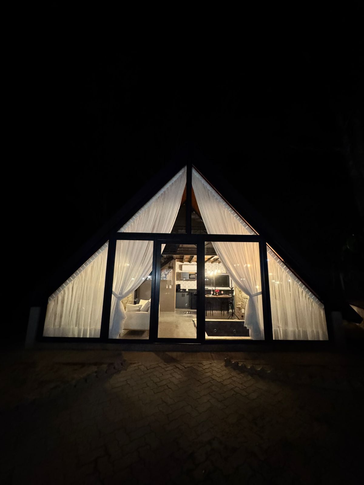 The exterior of a triangular-shaped chalet is visible, designed with large glass panels and sheer curtains. The warm glow of interior lighting softly illuminates the space, highlighting a welcoming atmosphere. The surrounding area remains dark, accentuating the structure's inviting presence.