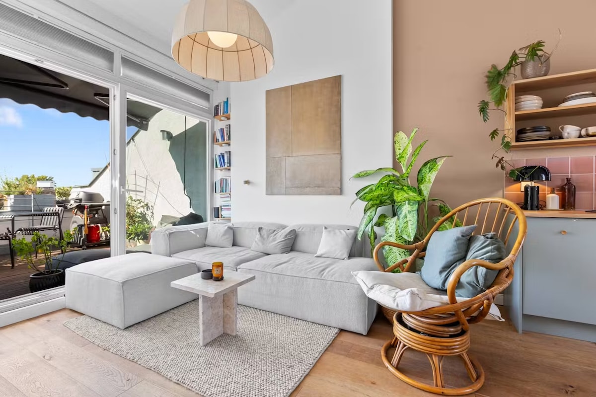 A bright living area features a light-colored sectional sofa paired with a minimalist coffee table. A rattan chair is positioned nearby, complemented by a vibrant green plant. Large glass doors provide access to the balcony, showcasing inviting outdoor seating.