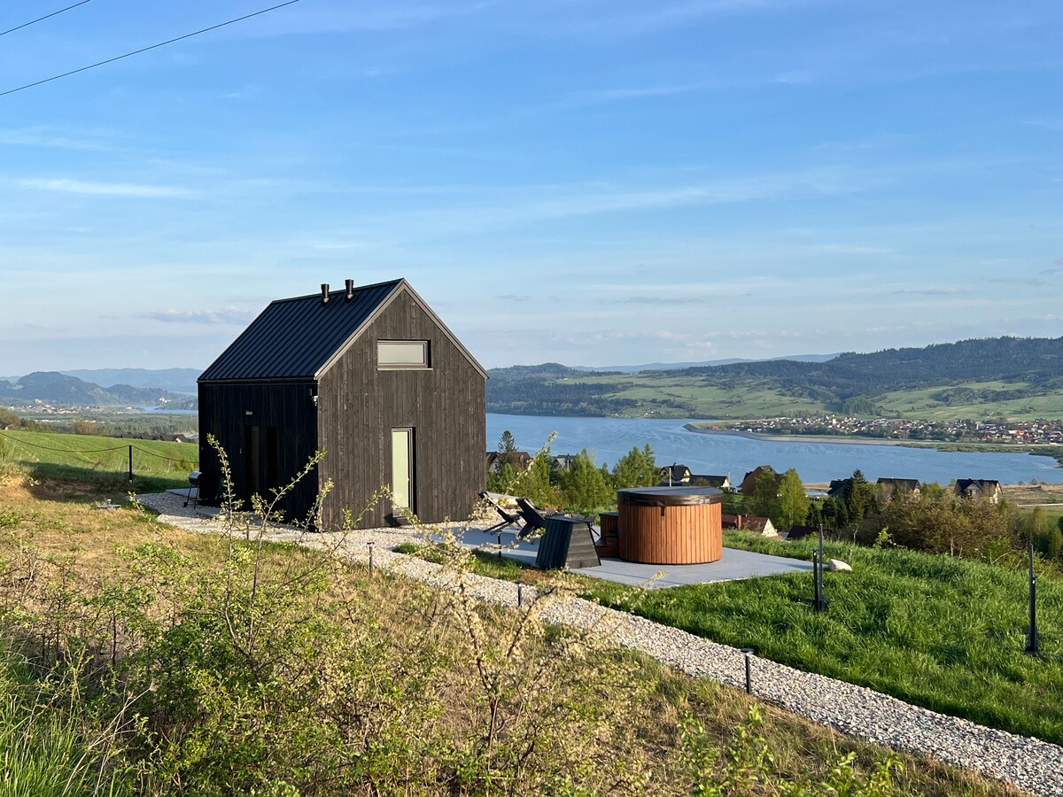 A modern wooden cabin is set against a backdrop of lush greenery and rolling hills. An outdoor hot tub is situated on a patio, offering scenic views of a nearby lake. Smooth stones create a path leading to the cabin, enhancing the serene setting.