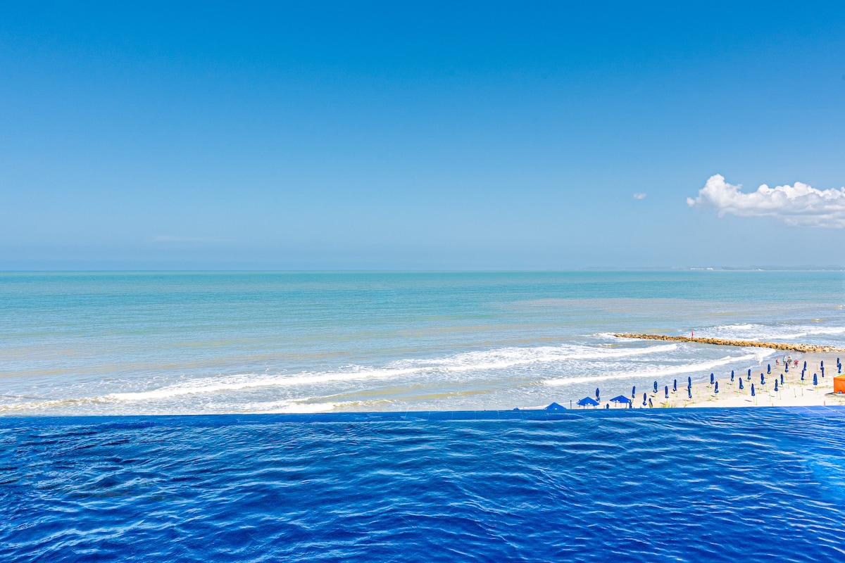 An expansive view of the ocean stretches to the horizon, showcasing shades of blue water meeting a clear sky. The beach is lined with umbrellas, while gentle waves create a serene atmosphere along the shore.