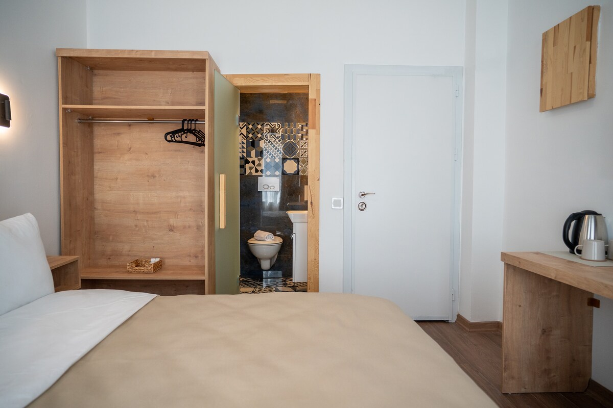 A modern room features a spacious bed with neutral-toned bedding. A wooden wardrobe is partially open, revealing hanging items. A compact bathroom is visible through a door, showing decorative tiles. A simple desk with a kettle is positioned nearby, enhancing the functionality of the space.