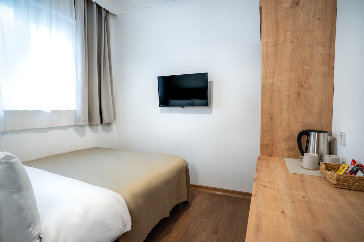 A modest guest room features a spacious bed with a neutral-colored coverlet. A wall-mounted television is positioned opposite the bed. A wooden cabinet holds a kettle and a basket, providing tea-making essentials. Natural light filters through sheer curtains, enhancing the room's simplicity.
