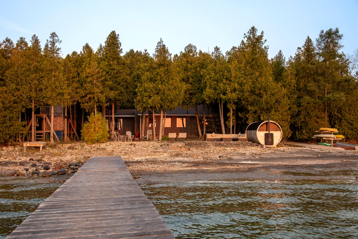 The cabin is nestled among trees along the shoreline, featuring a wooden dock leading to a pebble beach. Outdoor seating and a play structure are visible in the yard, while kayaks and a rowboat are stored nearby.