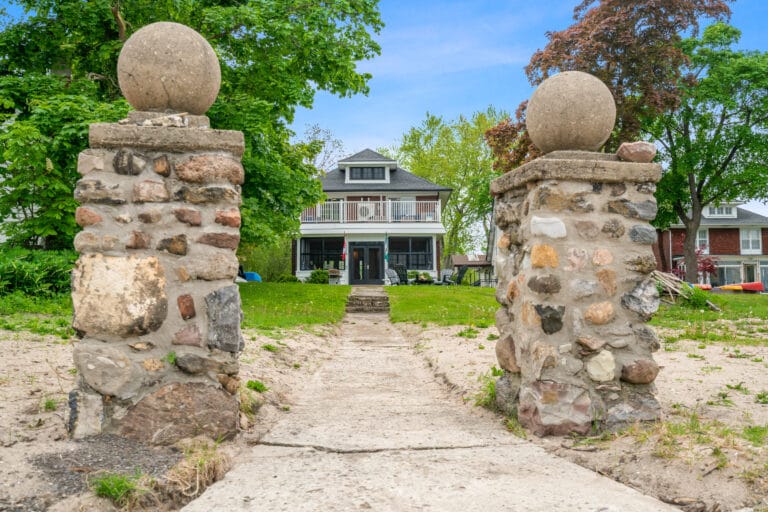 A stone pathway leads to a large house framed by lush greenery. Two round stone pillars mark the entrance, adding a touch of elegance. The home's exterior features a welcoming porch with railings, inviting guests to explore further.