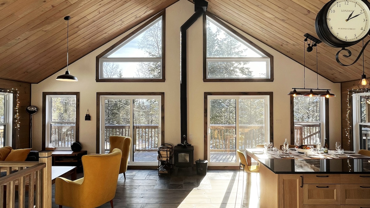 A spacious living area features a cathedral ceiling with large windows, allowing natural light to fill the room. A cozy wood stove is positioned near a modern kitchen island, accompanied by a dining table set for guests. Comfortable yellow chairs provide inviting seating.
