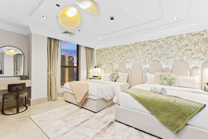 Un dormitorio luminoso con dos camas, papel pintado con motivos botánicos, cortinas beis y un tocador con espejo.