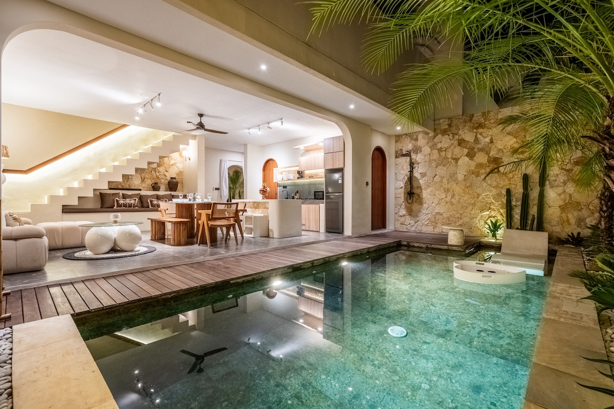 A serene view of the private pool, framed by tropical plants and stone walls. The inviting waters reflect the ambient lighting from the nearby kitchen and living area. Soft outdoor seating is positioned around the pool, creating a perfect space for relaxation and socializing.