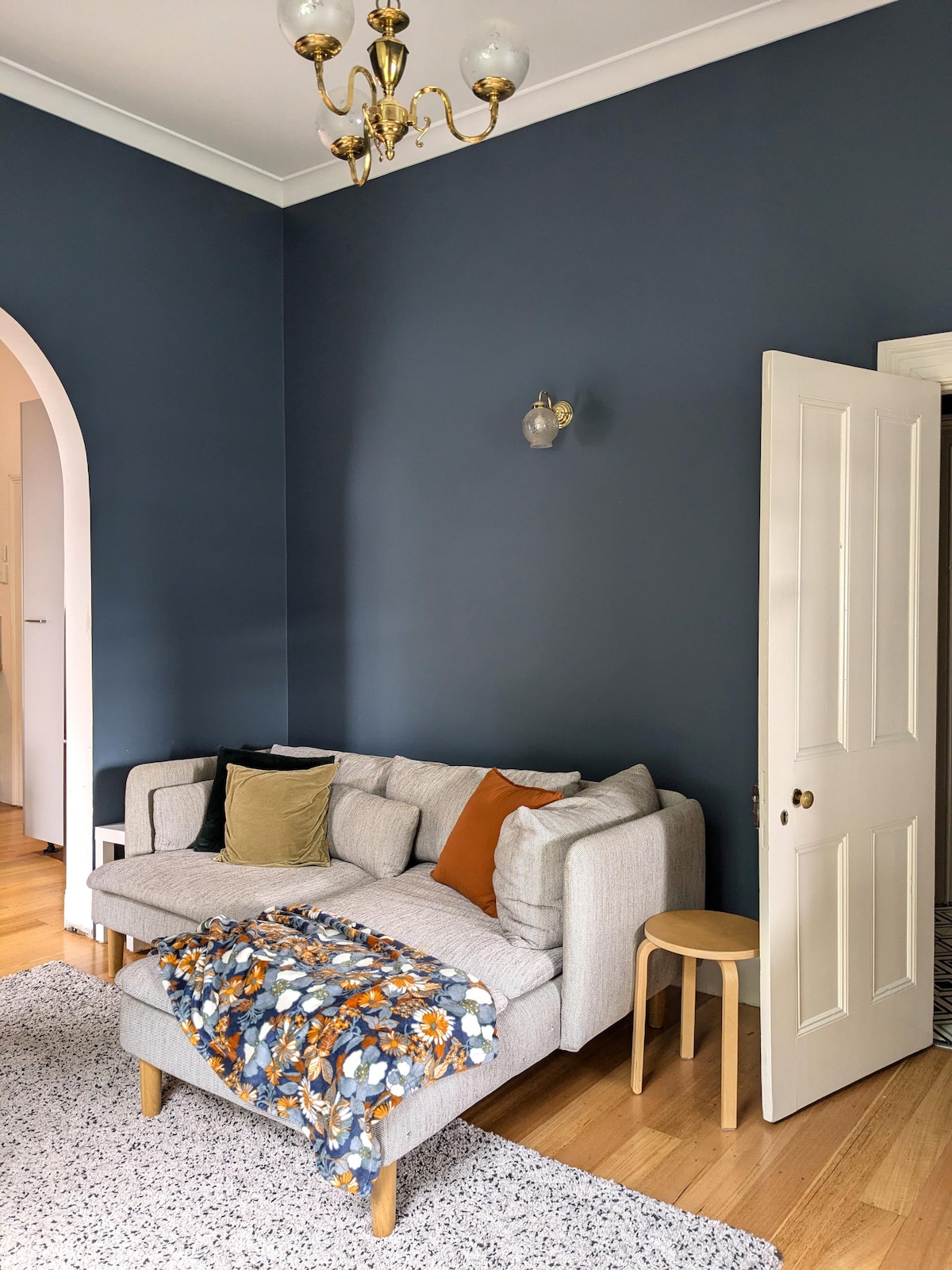 A light grey sectional sofa is positioned against a deep blue wall, featuring a cozy throw in floral patterns draped over its arm. A small wooden stool sits nearby, while a soft area rug covers the wooden floor. An archway leads to another room.